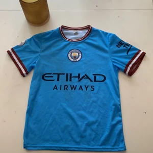 Blå Manchester City fotbollströja - Säljer en blå Manchester City fotbollströja med korta ärmar och klubbens emblem på bröstet. Tröjan har sponsorloggan 'Etihad Airways' på framsidan och spelarens namn och nummer 'De Bruyne 17' på baksidan. Perfekt för fans som vill visa sitt stöd!💙
