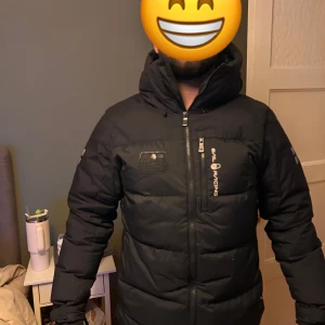 Svart pufferjacka från Sail Racing - Snygg svart pufferjacka från Sail Racing med dragkedja och huva. Jackan har långa ärmar och logotypdetaljer på bröstet och ärmen. Perfekt för kalla dagar.