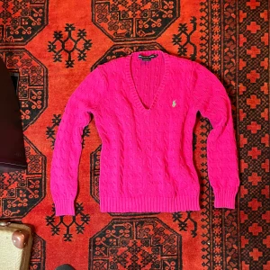 Rosa stickad tröja från Ralph Lauren - Säljer en snygg rosa stickad tröja från Ralph Lauren med kabelstickat mönster och en grön logga på bröstet. Tröjan har en v-ringad hals och långa ärmar. Perfekt för att liva upp garderoben!