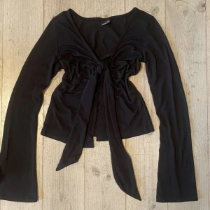 Svart långärmad topp med knyt - Snygg svart långärmad topp från Gina Tricot med knyt framtill. Perfekt för en stilren look. Toppen har en v-ringad design och är tillverkad i ett mjukt material.