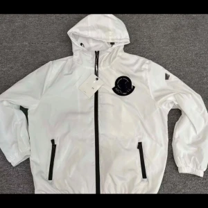 Vit vindjacka med huva - Säljer en stilren vit vindjacka med huva och svarta detaljer. Jackan har en dragkedja framtill och två fickor med dragkedjor. Perfekt för blåsiga dagar. Storlek Medium, använd ca 2 veckor. Säljer då jag vill bli av med allt hemma kan pruta snabb affär