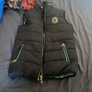 Svart dunväst från Jacson - Säljer en svart dunväst från Jacson med dragkedja och två fickor framtill. Västen har ett märke på bröstet och gröna detaljer vid dragkedjorna. Perfekt för ridning och kyligare dagar. En perfekt väst att ha i stallet nu mot våren fick den i födelsedagspresent och använde den mycket men den är fortfarande väldigt fin. Bra kvalitet då jag har använt den 3 vårar och höstar. 140 storlek.