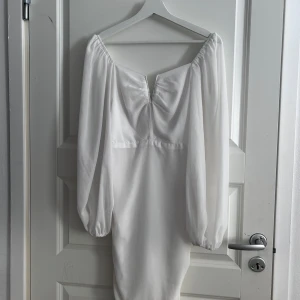 Vit klänning med v-ringning - Elegant vit klänning med v-ringad design och långa ärmar. Klänningen har en smickrande rynkad detalj på framsidan som ger en fin siluett. Perfekt för speciella tillfällen.