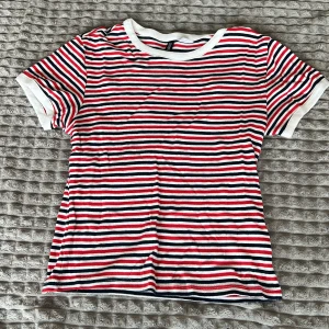 Randig t-shirt  - Säljer en randig t-shirt i bomull med röd, vit och blå färg. Den har korta ärmar och en klassisk rund halsringning. Perfekt för en avslappnad stil.
