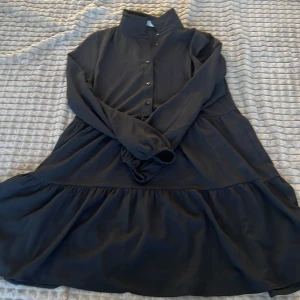 Svart klänning med knappar - Säljer en elegant svart klänning med knappar framtill och långa ärmar. Klänningen har en volangdetalj vid kjolen som ger en fin rörelse. Perfekt för både vardag och festliga tillfällen.