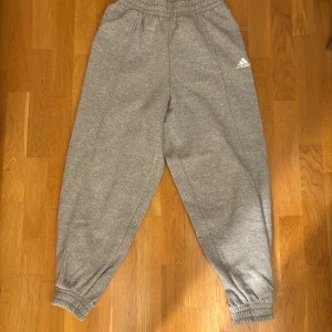 Grå mjukisbyxor från Adidas - Säljer ett par grå mjukisbyxor från Adidas med elastisk midja och muddar vid bensluten. Perfekta för en avslappnad stil. Adidas-loggan är tryckt på ena benet. Superbekväma och stilrena!
