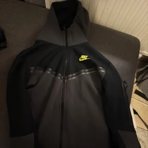 Nike tech fleece  - Säljer en snygg svart och grå hoodie från Nike med dragkedja och huva. Jackan har en gul Nike-logga på bröstet och praktiska fickor med dragkedja. Perfekt för en sportig look! Storlek S
