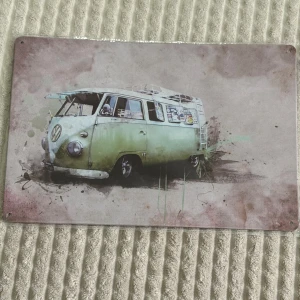 Vintage plåtskylt med VW-bussmotiv - Säljer en retroinspirerad plåtskylt med motiv av en klassisk VW-buss i grönt och beige. Skylten har en sliten look med färgstänk och är perfekt för att ge en nostalgisk känsla till ditt rum eller garage. Den har förborrade hål i hörnen för enkel upphängning.