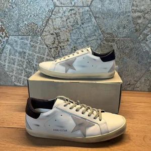 Golden Goose superstar - Säljer ett par stilrena vita sneakers från Golden Goose med en grå stjärna på sidan och svart häl. Skorna har snörning och en klassisk design med en touch av lyx. Perfekta för en avslappnad men trendig look. Skorna är helt oanvända och därmed i perfekt skick! I köpet ingår box och dustbag. Nypris på dessa ligger runt 6000 medan mitt pris är 2800! De är självklart äkta och ifall ni har frågor är det endast att skriva till mig!