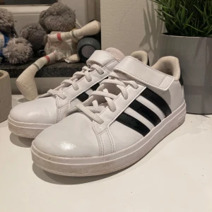Vita sneakers från Adidas - Säljer ett par vita sneakers från Adidas med klassiska svarta ränder. Skorna har både snörning och kardborreband för extra komfort och stil. Perfekta för en avslappnad look. Använda max 2 gånger. Säljer för de var för stora!