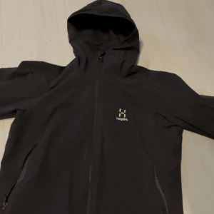 Svart vindjacka från Haglöfs - Säljer en svart vindjacka från Haglöfs med huva och dragkedja framtill. Jackan har en stilren design med två fickor med dragkedjor på sidorna. Perfekt för blåsiga dagar.