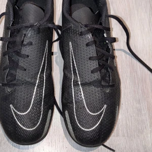 Svarta fotbollsskor från Nike - Snygga svarta fotbollsskor från Nike med vita detaljer och snörning. Perfekta för spel på gräs med sina dobbar som ger bra grepp. Skorna har en modern design med Nike-loggan på sidan.
