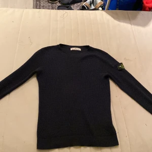 Svart stickad tröja från Stone Island - Säljer en stilren svart stickad tröja från Stone Island. Tröjan har långa ärmar och en klassisk rund halsringning. Perfekt för kyligare dagar och ger en snygg look med den ikoniska loggan på ärmen.