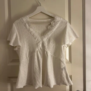 Vit blus med spetsdetaljer - Charmig vit blus med v-ringning och spetsdetaljer. Blusen har korta ärmar och ett fint hålmönster som ger en romantisk känsla. Perfekt för varmare dagar.