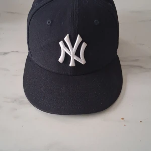 Svart keps från New Era - Snygg svart keps från New Era med den ikoniska NY-loggan broderad i vitt framtill. Kepsen är en del av 59FIFTY-serien och har en klassisk passform. Perfekt för att ge din outfit en sportig touch.
