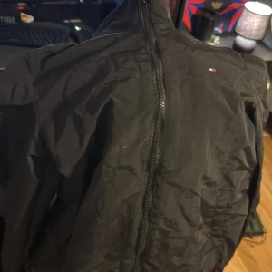 Svart vindjacka från Tommy Hilfiger - Säljer en stilren svart vindjacka från Tommy Hilfiger. Jackan har en dragkedja framtill och det ikoniska logotypmärket på bröstet. Perfekt för blåsiga dagar och en snygg detalj i garderoben.