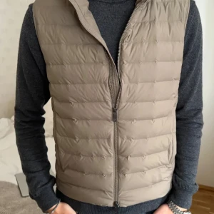 Beige dunväst med dragkedja - Snygg beige dunväst med dragkedja framtill. Perfekt för lager-på-lager under kyliga dagar. Västen har en stilren design och passar bra till både casual och mer uppklädda tillfällen.