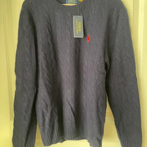 Blå kabelstickad tröja från Polo Ralph Lauren - Säljer en stilren grå kabelstickad tröja från Polo Ralph Lauren. Tröjan har en klassisk rund halsringning och det ikoniska röda logotypbroderiet på bröstet. Perfekt för en elegant och avslappnad look. Nypris - 2 695