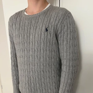 Grå stickad tröja från Ralph Lauren - Säljer en stilren grå stickad tröja från Ralph Lauren med kabelstickat mönster. Tröjan har långa ärmar och en rund halsringning. Perfekt för kyligare dagar.