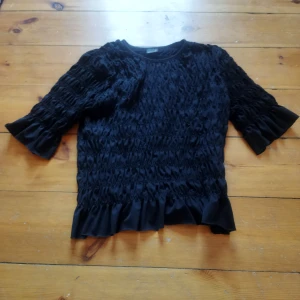 Svart topp från Gina Tricot - Snygg svart topp från Gina Tricot med ett unikt rynkat mönster och volangdetaljer vid ärmslut och nederkant. Perfekt för att ge din outfit en elegant touch.