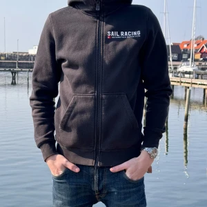 Svart hoodie från Sail Racing - Säljer en svart hoodie från Sail Racing med dragkedja och tryck på både fram- och baksidan. Tröjan har långa ärmar och en praktisk huva. Perfekt för kyliga dagar vid vattnet eller en avslappnad dag på stan.