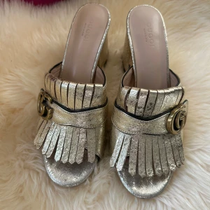 Guldiga klackskor från Gucci - Snygga guldiga klackskor från Gucci med fransdetaljer och en elegant GG-logga. Perfekta för att ge en lyxig touch till din outfit. Skorna har en öppen tå och en bekväm klack.