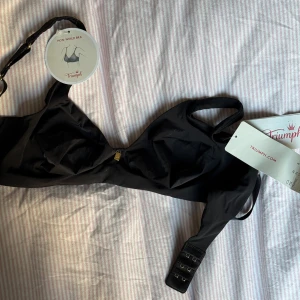 Svart BH från Triumph - Säljer en elegant svart BH från Triumph utan bygel. Den har en liten gulddetalj framtill och justerbara axelband för bästa passform. Perfekt för en bekväm och stilren look.