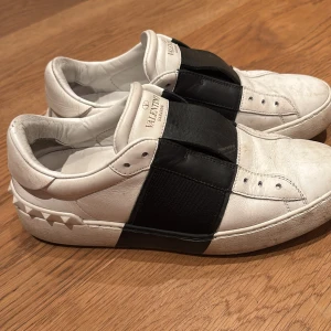 Valentino Open sneakers i vitt och svart 36 - Snygga Valentino Open sneakers i vitt läder med en bred svart rem över vristen. Skorna har de ikoniska nitarna på hälen och en stilren design. Perfekta för en trendig look. Skick 8/10. Storlek 36