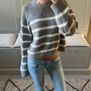 Grå och vit randig stickad tröja - Säljer en mysig grå och vit randig stickad tröja med croppad passform. Tröjan har långa ärmar och en avslappnad stil, perfekt för kyligare dagar. Den är både stilren och bekväm, ett måste i garderoben!