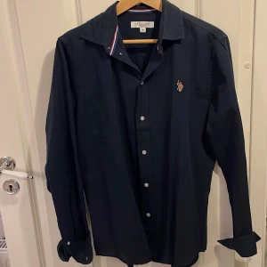 Mörkblå skjorta från U.S. Polo Assn. - Snygg mörkblå skjorta från U.S. Polo Assn. med broderad logga på bröstet. Skjortan har långa ärmar och knappar framtill. Perfekt för en stilren look.