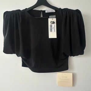 Svart topp från ADOORE - Snygg svart topp från ADOORE med puffiga ärmar och en elegant design. Perfekt för att ge en stilren look. Toppen har en rund hals och en kortare passform.