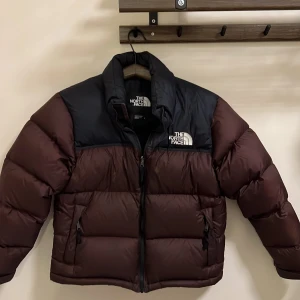 The North Face - dunjacka  - Säljer en snygg brun och svart dunjacka från The North Face. Jackan har en dragkedja framtill och är perfekt för kalla dagar. Den är långärmad och har en klassisk pufferdesign med märkets logga på bröstet.