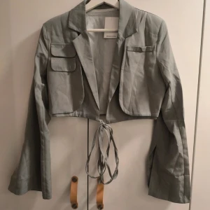 Grå croppad kavaj med knytband - Snygg grå croppad kavaj från Junkies Clothing med knytband i midjan. Kavajen har långa ärmar och en stilren design med fickdetaljer på framsidan. Perfekt för en trendig look!