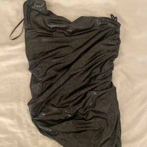 Svart glittrig one shoulder-topp från H&M Divided - Säljer en svart glittrig one shoulder-topp från H&M Divided. Den har en tight passform och är tillverkad i ett glittrigt syntetmaterial. Perfekt för dig som vill sticka ut med en trendig och elegant look.