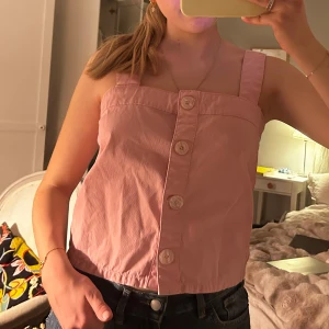 Rosa linnetopp med knappar - Söt rosa linnetopp med breda axelband och stora dekorativa knappar framtill. Toppen har en rak passform och är tillverkad i ett lätt bomullsmaterial, perfekt för varma dagar.