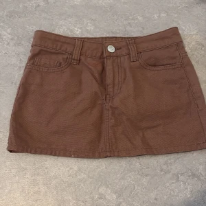 Brun minikjol i jeansmaterial - Säljer en brun minikjol i jeansmaterial med klassiska fickor fram och bak samt knappstängning. Kjolen har bälteshällor och en enkel, stilren design som passar till många olika looks.