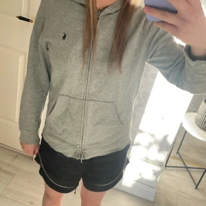 Grå hoodie från Polo Ralph Lauren - Säljer en grå hoodie från Polo Ralph Lauren med dragkedja och två fickor framtill. Klassisk marinblå logga på bröstet och mjukt bomullsmaterial. Perfekt som en extra tröja till sommaren