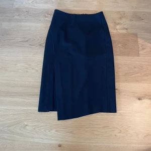 Mörkblå pennkjol från Dagmar - Säljer en stilren svart pennkjol från Dagmar med hög midja och slits på sidan. Kjolen har en rak passform och stängs med dragkedja bak. Perfekt för dig som gillar en klassisk och elegant look.