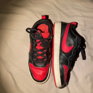Nike Dunk Low svart/röd - Säljer ett par Nike Dunk Low i svart och rött med klassisk röd sula och svart snörning. Skorna har en låg siluett, rund tå och tydlig Nike-logga på hälen. Perfekta för dig som gillar streetwear och vill sticka ut med färg.