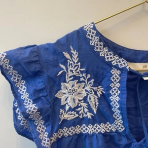 Blå blus med broderi från H&M - Supersöt blå blus från H&M med vita broderade blommor och dekorativa band med tofsar framtill. Blusen har korta ärmar och är gjord i ett lätt bomullsmaterial. Perfekt för en somrig look!