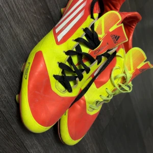 Adidas F30 fotbollsskor i röd och gul - Säljer ett par Adidas F30 fotbollsskor i en riktigt snygg röd och gul färgkombination med svarta snören och vita detaljer. Perfekta för dig som vill sticka ut på planen!