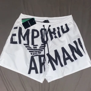 Vita badshorts från Emporio Armani - Snygga vita badshorts från Emporio Armani med stor svart logga och text framtill. Shortsen har snörning i midjan och elastisk resår. Perfekta för stranden eller poolen, och levereras i en praktisk förvaringspåse.