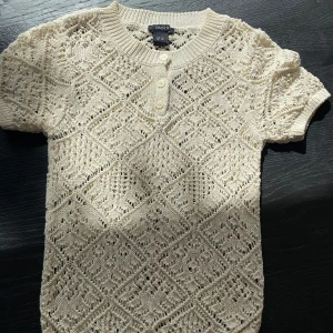 Beige stickad topp från Lindex - Säljer en beige stickad topp från Lindex med korta ärmar och v-ringning med tre knappar framtill. Tröjan har ett fint hålmönster och är perfekt för vår och sommar. Superfin att styla till både jeans och kjol! Är använd 1 gång☺️