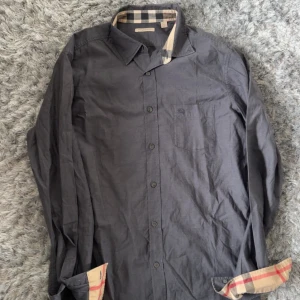 Mörkgrå skjorta från Burberry - Säljer en mörkgrå skjorta från Burberry med klassiskt rutigt tyg på insidan av kragen och ärmsluten. Skjortan har knappar framtill, bröstficka och är långärmad. Perfekt för dig som gillar stilrena plagg med en twist.