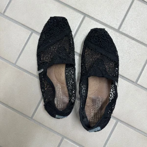 Svarta spetsballerinaskor från TOMS - Säljer ett par svarta ballerinaskor från TOMS med vacker spets och rund tå. Skorna har en lätt och luftig design med TOMS-logga bak på hälen. Använda cirka 5 gånger💗