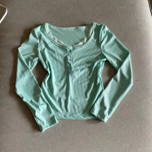 Ljusblå ribbad långärmad topp med spetsdetalj - Säljer en ljusblå ribbad långärmad topp med rund halsringning och söt vit spetskant. Toppen har knappar framtill och är croppad i modellen. Perfekt för en chill och stilren look.