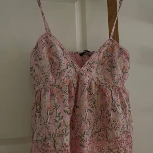 Rosa blommig topp från BikBok - Supersöt rosa topp från BikBok med smala axelband och v-ringning. Toppen  har blommigt mönster, broderade detaljer och spetskant nertill. Perfekt för sommarens alla tillfällen!