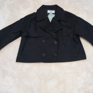Svart kort trenchcoat från H&M - Säljer en svart kort trenchcoat från H&M i storlek 146 (10-11 år). Jackan har dubbelknäppning framtill, bred krage och långa ärmar. Perfekt för dig som vill ha en stilren och klassisk look.