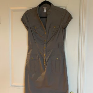 Beige skjortklänning från H&M - Stilren khakigrön skjortklänning från H&M med korta ärmar och knappar framtill i guldton. Klänningen har fickor med lock på bröstet och höfterna samt markerad midja. Perfekt för en avslappnad men snygg look.Använd en gång. Stl 38.