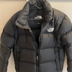 Svart dunjacka från The North Face - Säljer en svart dunjacka från The North Face med hög krage och dragkedja. Jackan har klassisk pufferstil och logga på bröstet. Perfekt för kalla dagar och riktigt skön att bära.